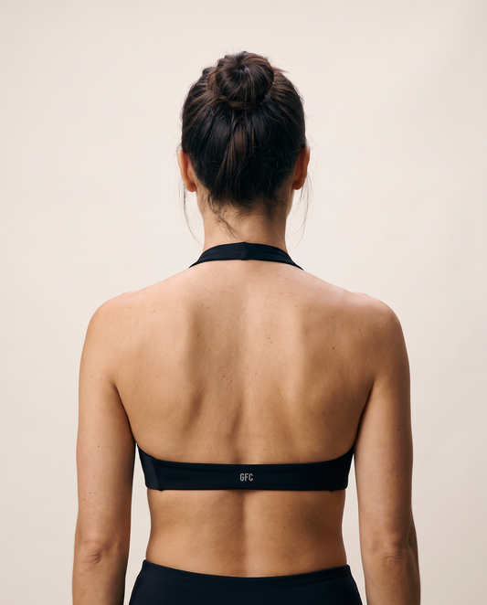 Rear view of a woman wearing a black halter-neck sports bra with a wide underband, shown against a plain beige background. The brand name 'BFC' is printed in white on the center of the band.