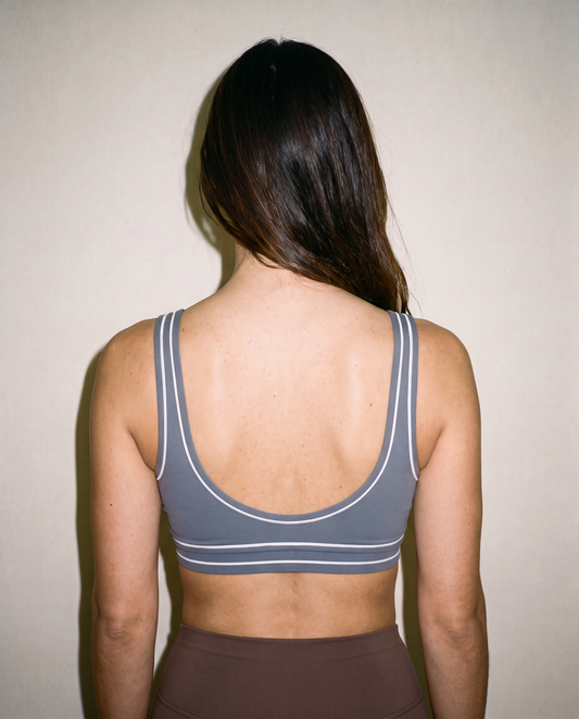 A woman viewed from behind wearing a gray sports bra with white stripe details, set against a plain beige wall.