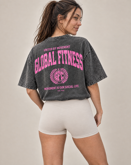 A woman wearing a faded charcoal gray oversized t-shirt with a bright pink 'GLOBAL FITNESS' graphic on the back, paired with white fitted shorts, shown from a three-quarters back view against a neutral beige background.