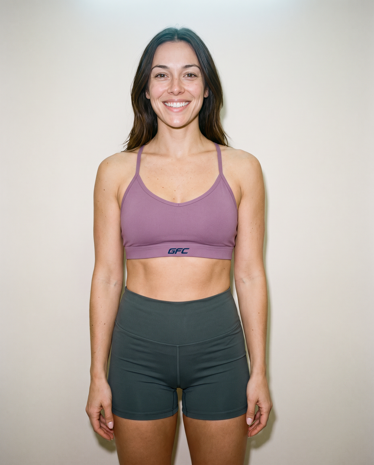 A woman modeling a mauve-purple sports bra and dark gray athletic shorts against a plain off-white wall. The sports bra features a small navy blue 'GFC' logo on the front band.