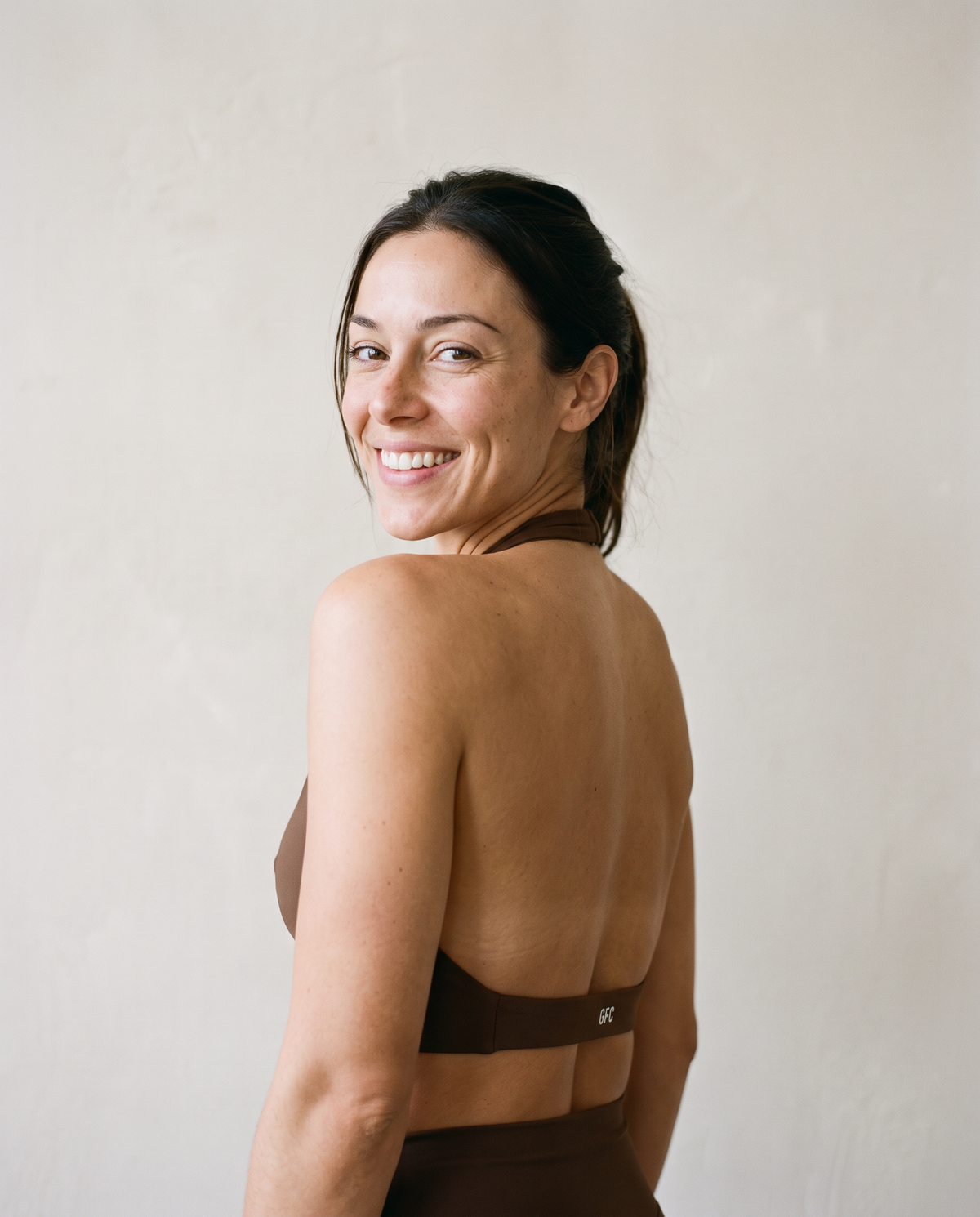 A woman with dark hair pulled back, seen from behind, wearing a brown athletic halter top and matching bottoms against a neutral, textured off-white wall.