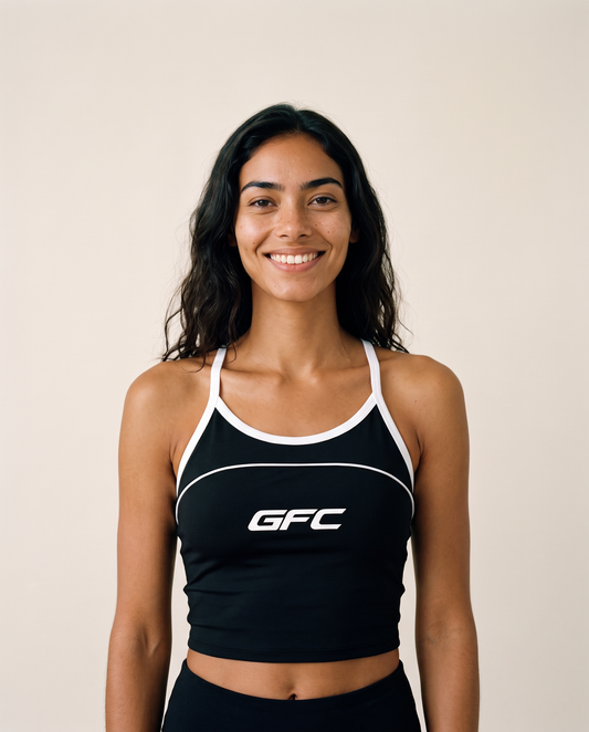 A smiling woman with long dark hair wearing a black athletic crop top with white trim and a white GFC logo on the chest, set against a plain beige background.