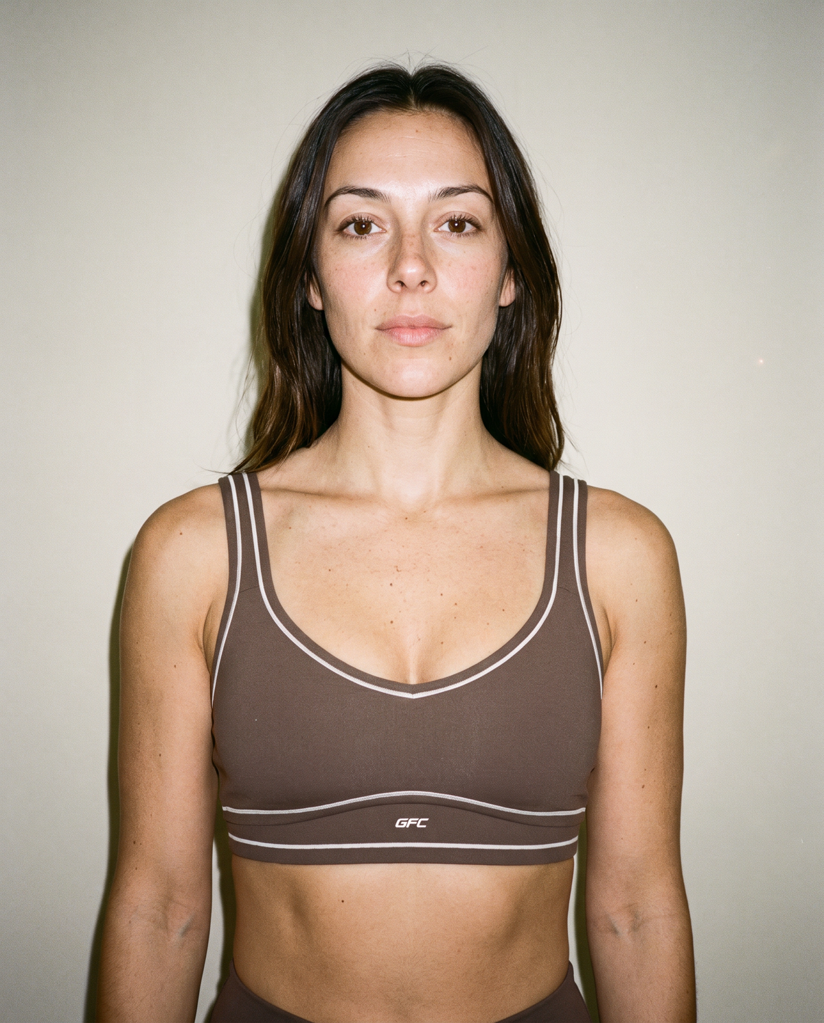 A woman wearing a brown sports bra with white piping and a small white GFC logo on the underband, photographed against a plain light beige wall.