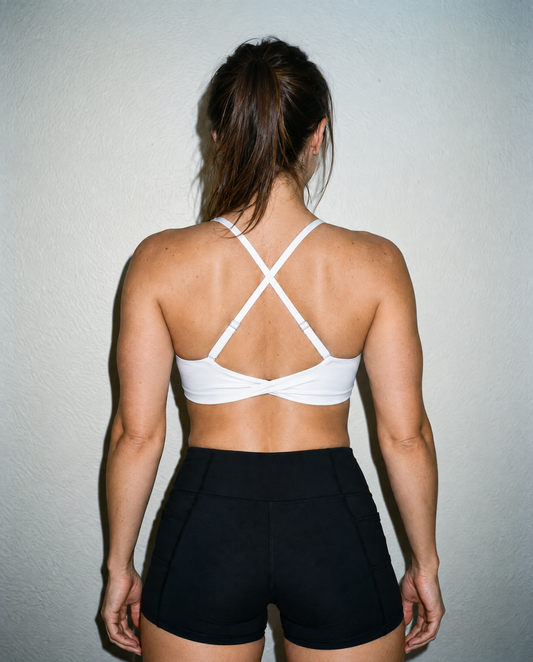 A woman viewed from behind wearing a white sports bra with crisscross back straps and black athletic shorts, standing against a plain light gray wall.
