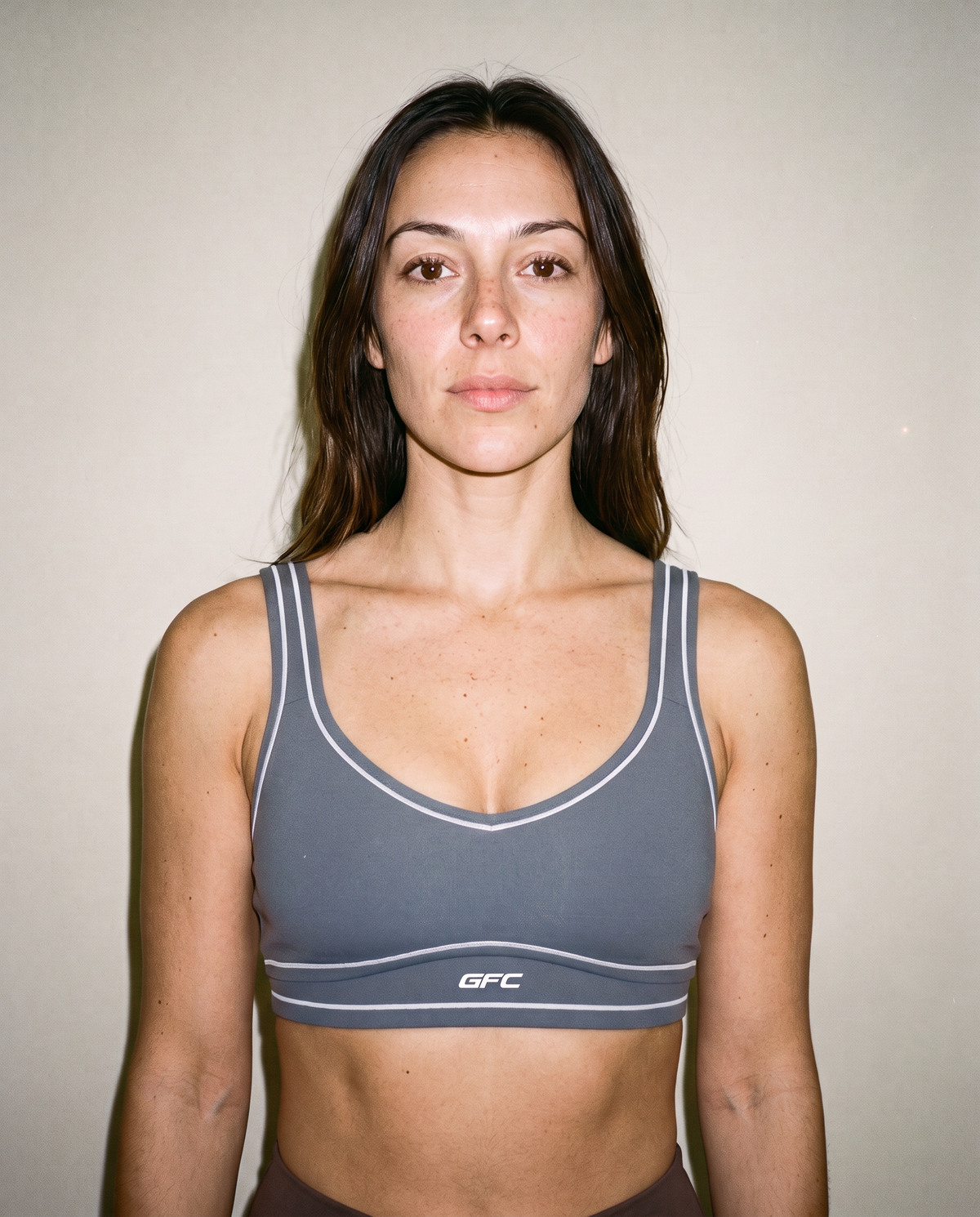 A woman wearing a gray sports bra with white piping and a white 'GFC' logo on the band, photographed against a plain beige wall.