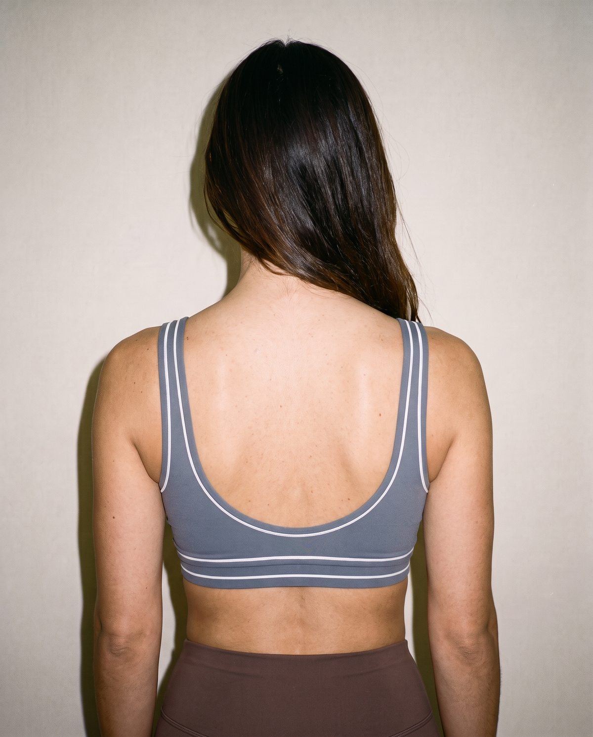 A woman viewed from behind wearing a gray sports bra with white stripe details, set against a plain beige wall.