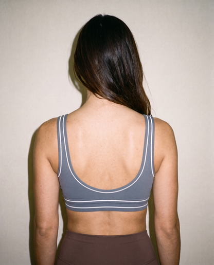 A woman viewed from behind wearing a gray sports bra with white stripe details, set against a plain beige wall.